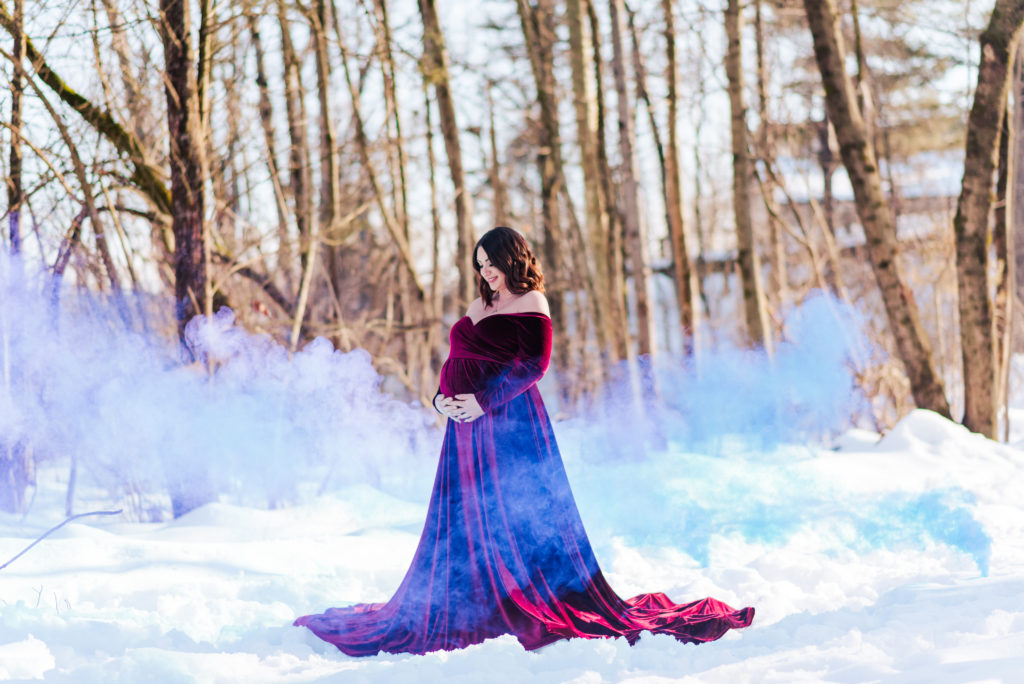 A mom-to-be in maroon velvet gown stands in the snow, with blue smoke surrounding her.