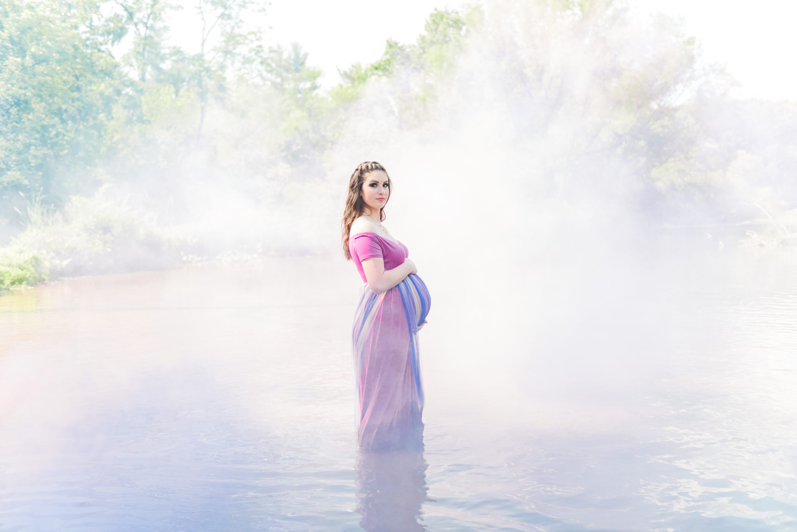 A mom-to-be in a oink and rainbow dress stands in a creek surrounded by colorful smoke